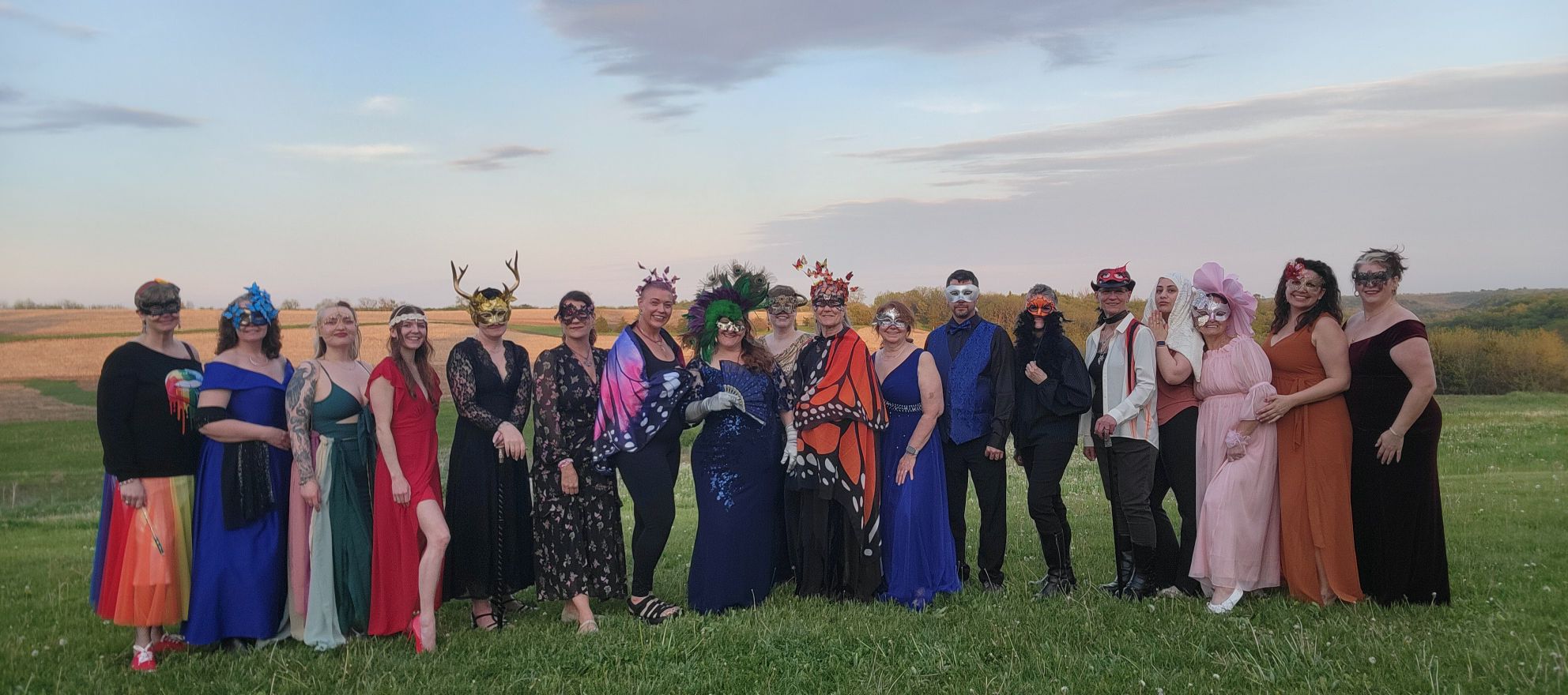Group of people in colorful masquerade costumes posing in a grassy field at sunset.